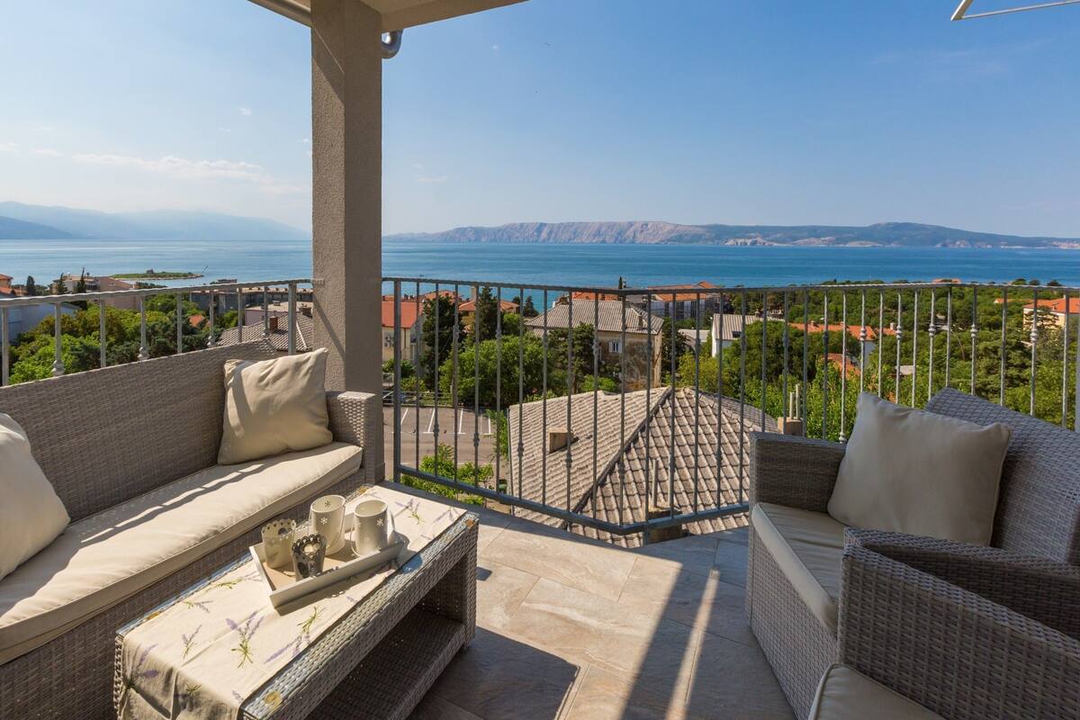 A balcony with wicker furniture, a tray with cups and a candle, and a view of the sea and buildings.