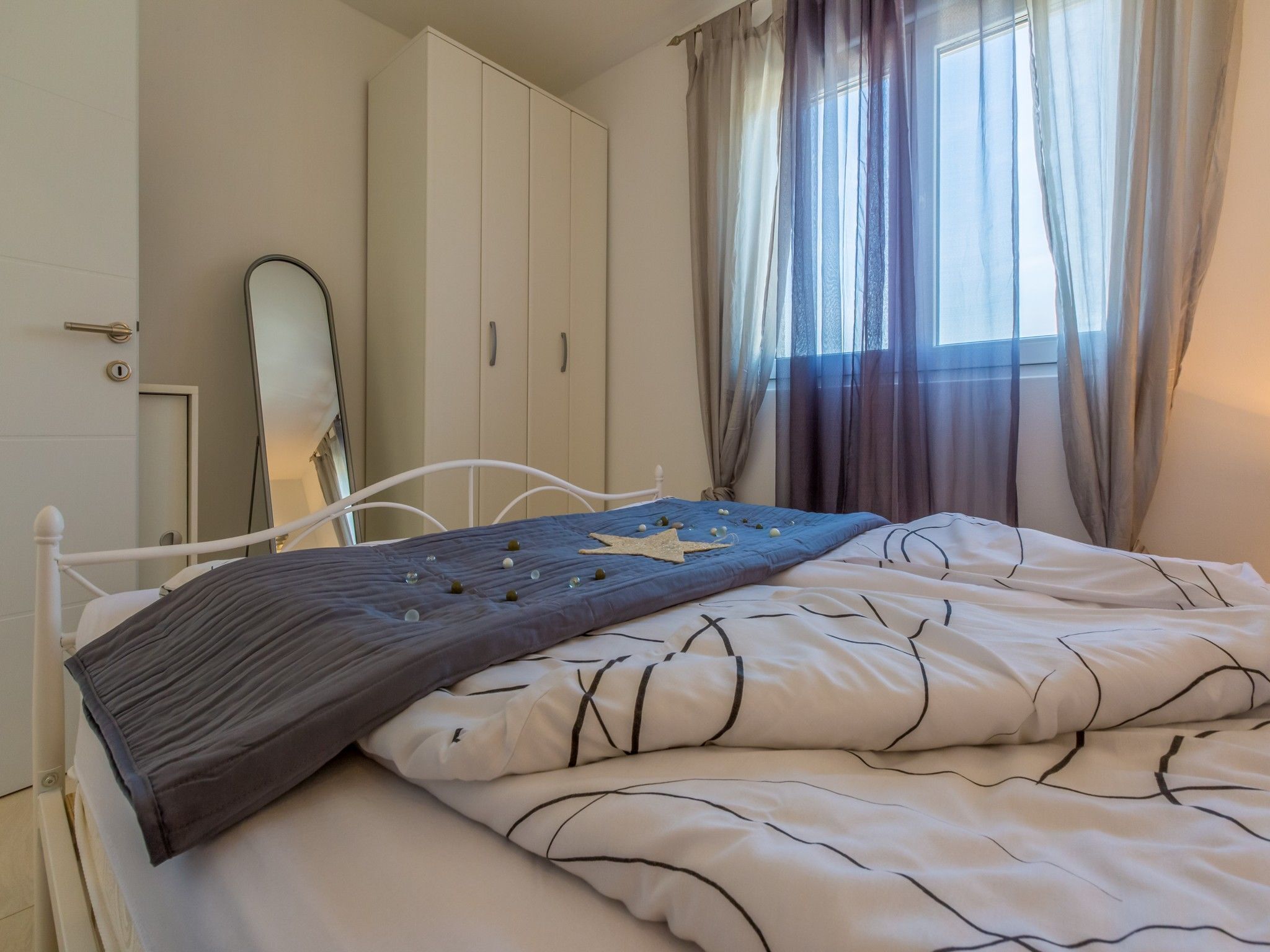 A neatly made bed with a blue blanket and white bedding, a white wardrobe, and a window with sheer curtains.