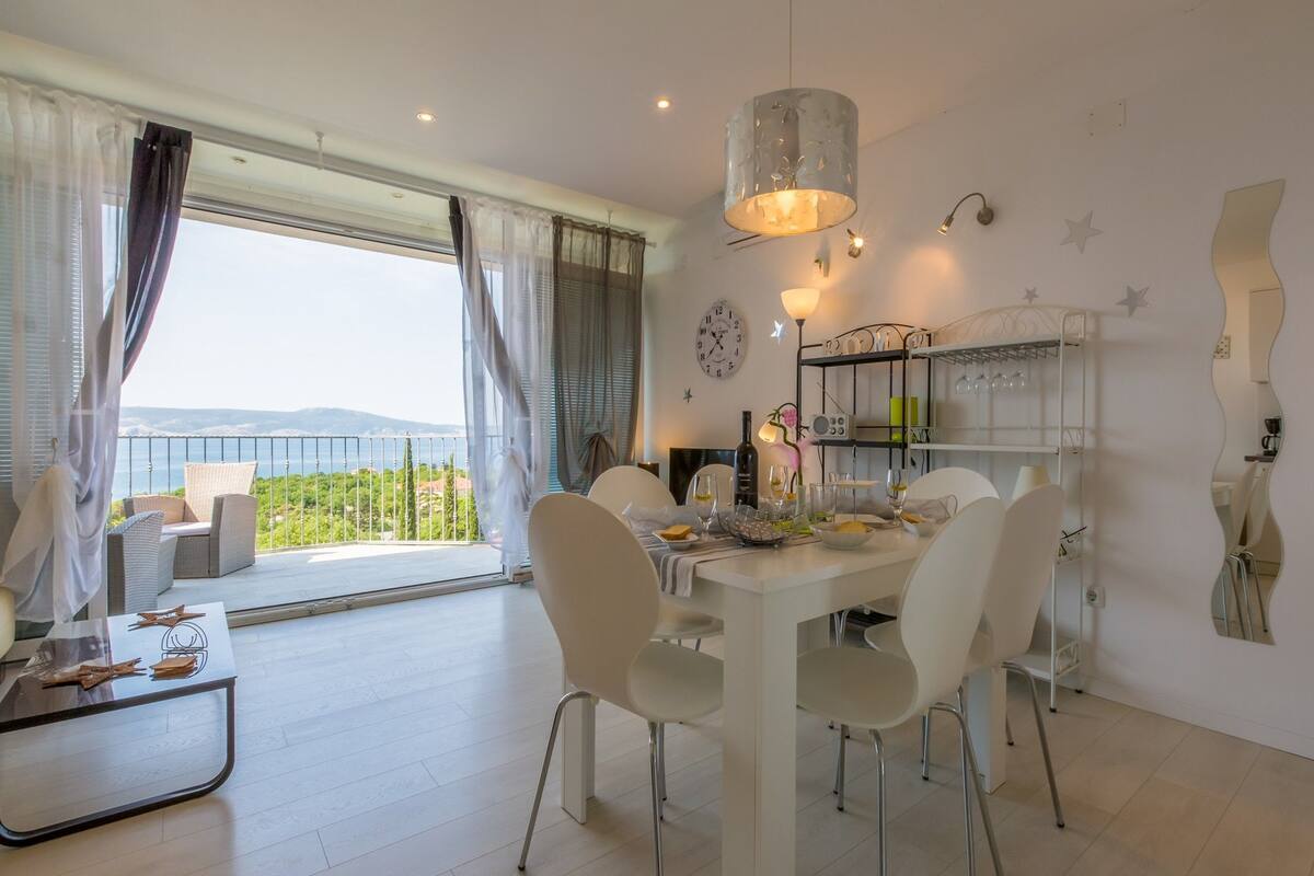 A dining area with a table set for four, a balcony with wicker furniture, and a view of the sea.