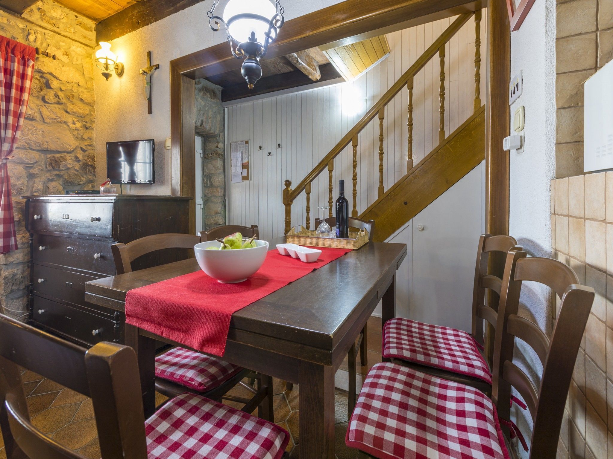 A dining area with a wooden table, checkered chairs, a red table runner, and a staircase in the background.