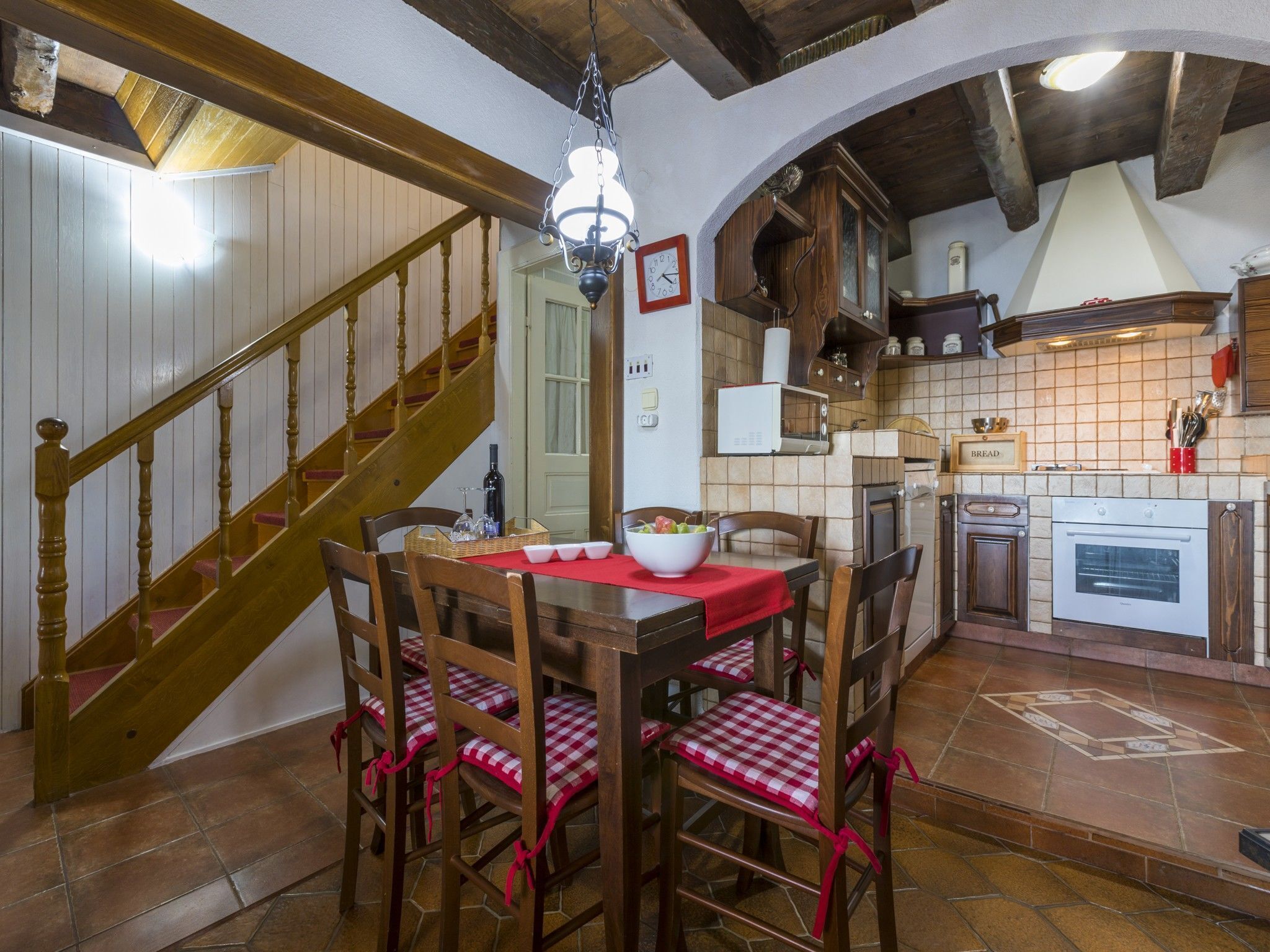 A dining area with a wooden table and chairs, a chandelier, and a kitchen in the background.