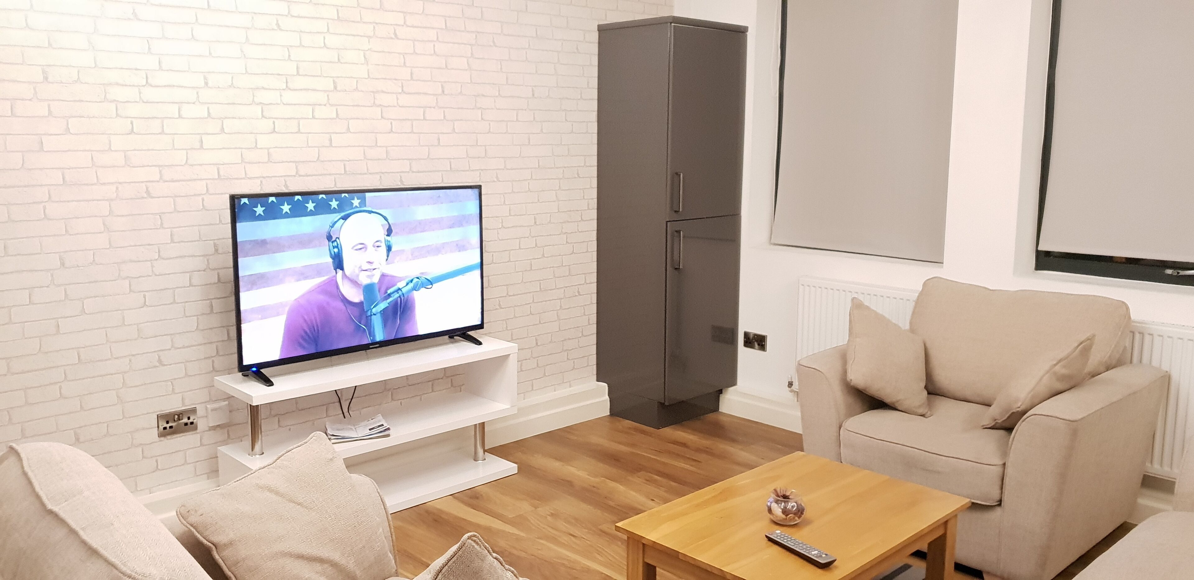 Un salon avec un téléviseur fixé à un mur en briques blanches, un meuble gris, un canapé beige et une table basse en bois.