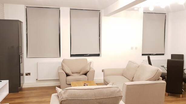 A modern living room with a sofa, armchairs, and a coffee table. There is a television on a stand, a radiator, and a window with blinds. - Manhattan 5 (Maidstone)
