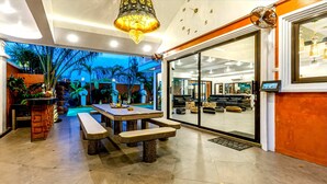 Um interior moderno com área de jantar, mesa e bancos, uma grande porta de vidro deslizante que dá para uma sala de estar, e teto com iluminação embutida e um lustre decorativo.