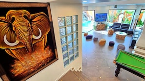 Uma mesa de bilhar em um ambiente com uma grande pintura de elefante, uma televisão e uma área de estar com pufes.
