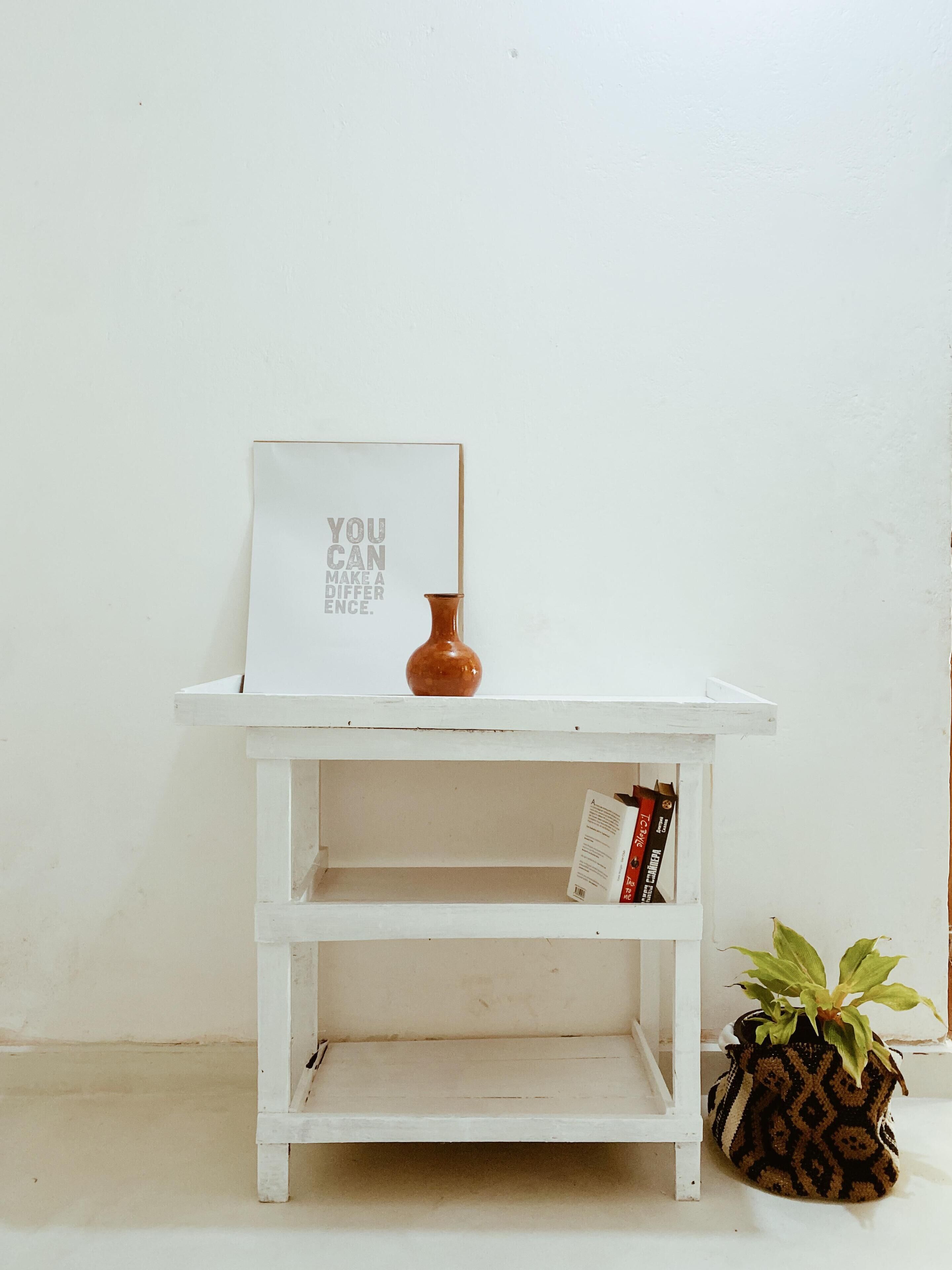 A white wooden shelf with a framed inspirational quote, a small brown vase, a book, and a potted plant.
