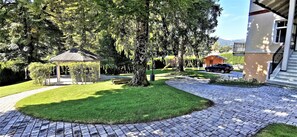 A paved pathway leading to a gazebo surrounded by lush greenery and a house with a visible staircase.