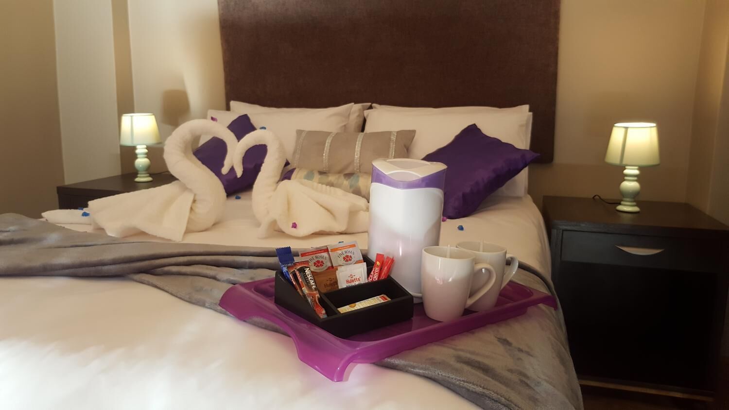 Una cama bien hecha con una bandeja morada que sostiene una taza, una caja de bombones y una caja de pañuelos.