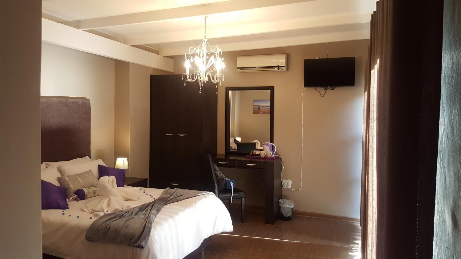 A hotel room with a bed, a desk, a chair, a chandelier, an air conditioning unit, and a television.