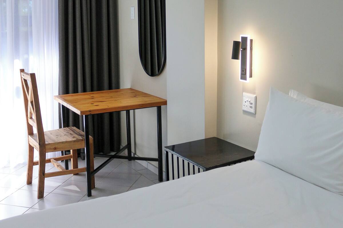 Hotellihuone, jossa on sänky, puinen tuoli, pöytä, seinään kiinnitetty valaisin ja ikkuna, jossa on verhot.