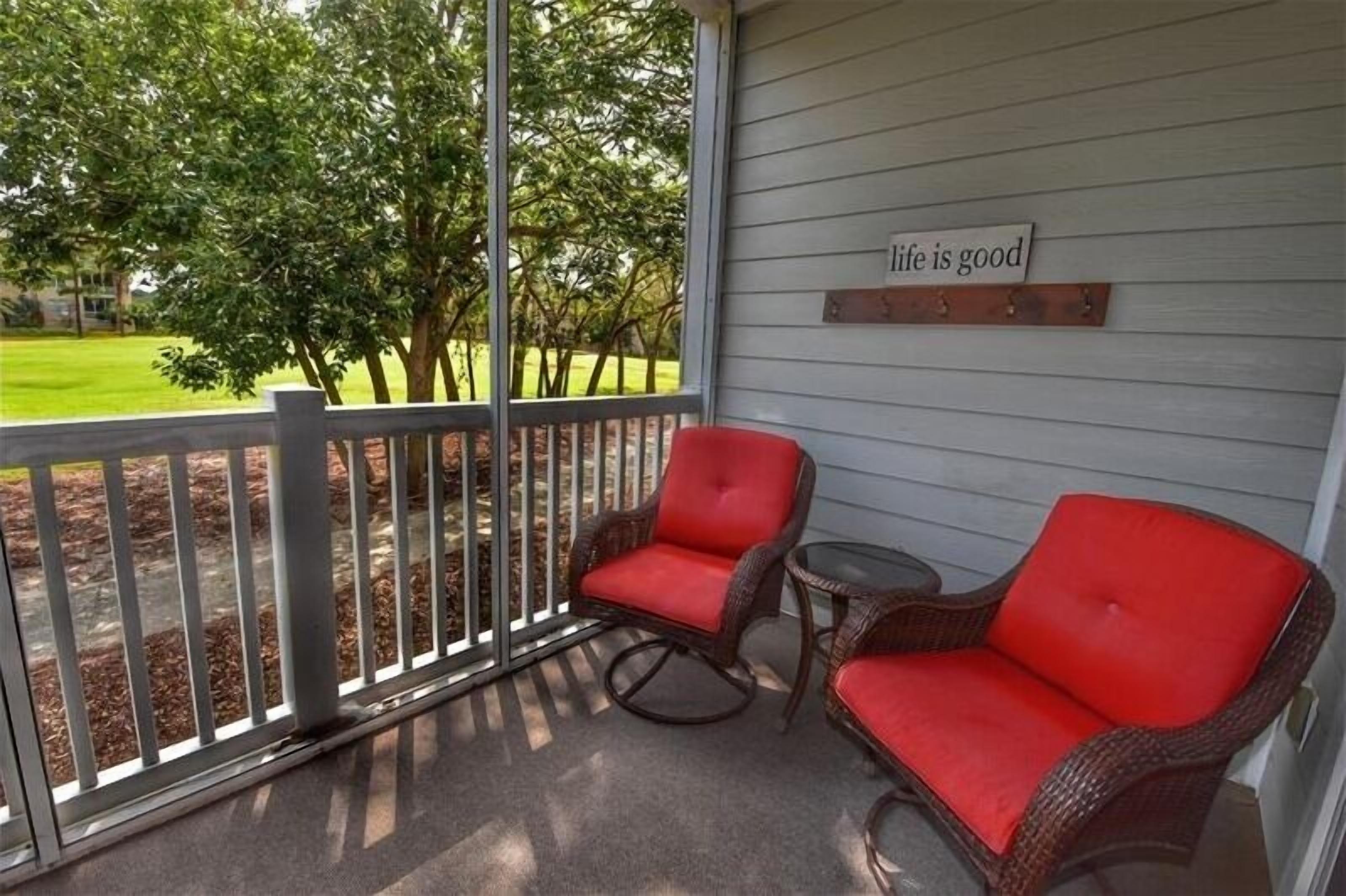 A covered porch with wicker chairs and a small table, a wooden sign on the wall, and a view of a green lawn and trees.