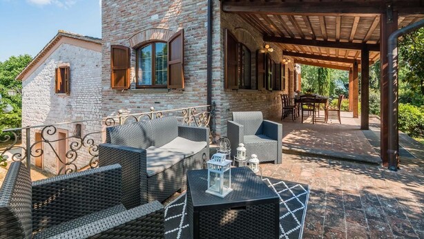 A covered outdoor seating area with wicker furniture and a table, adjacent to a stone building with wooden shutters.