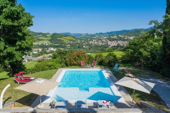 A swimming pool with lounge chairs and umbrellas, surrounded by greenery and a view of a town in the distance. - Villa Durante - Private villa, 5 bedrooms, pool, wifi, pets, Marche (Cagli)