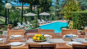 Un área de comedor junto a la piscina con mesas de madera dispuestas para una comida, rodeada de sillones de descanso y sombrillas.