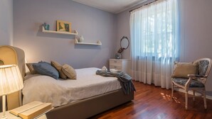 Un dormitorio con cama, mesita de noche, lámpara, silla y una ventana con cortinas traslúcidas.