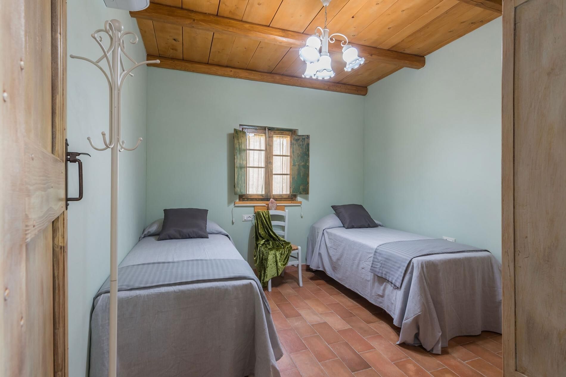 A small, cozy room with two single beds, a wooden ceiling, and a window with shutters.