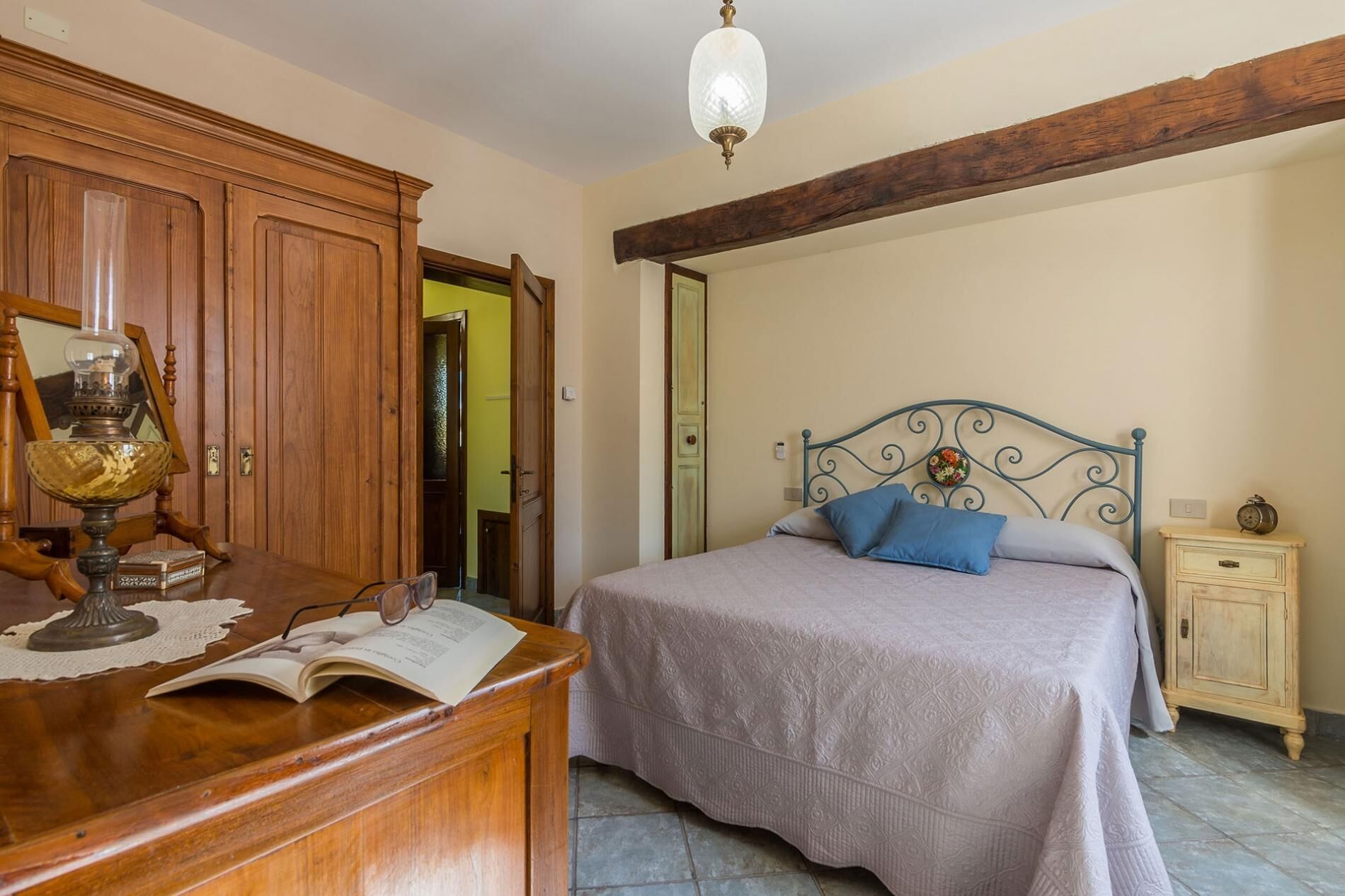 A bedroom with a wooden desk, an open book, a bed with a metal headboard, and a small bedside table with a clock.