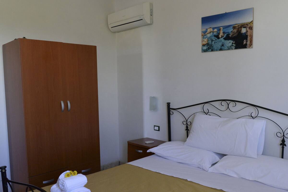 A bedroom with a bed, a wooden wardrobe, an air conditioner, a bedside table, and a painting on the wall.