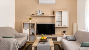 Una sala de estar con un televisor de pantalla plana, dos sofás, una mesa de centro con fruta, un reloj y un aparador con puertas de vidrio.