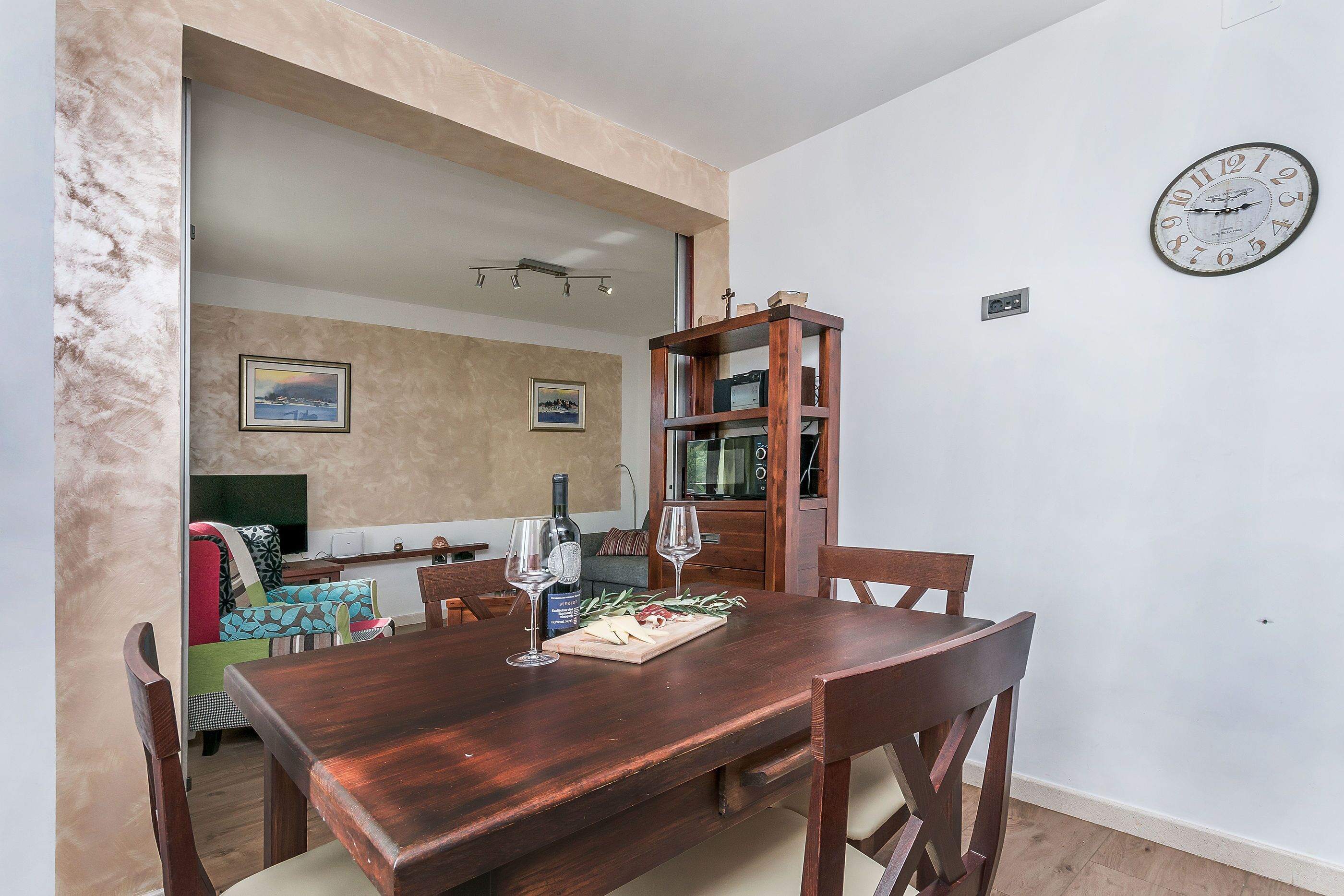 A dining area with a wooden table, chairs, a clock on the wall, and a bookshelf.