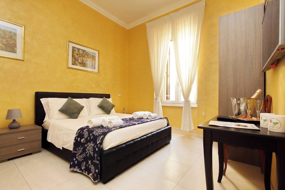 A neatly made bed with white linens and a dark headboard, a bedside table with a lamp, and a window with curtains.
