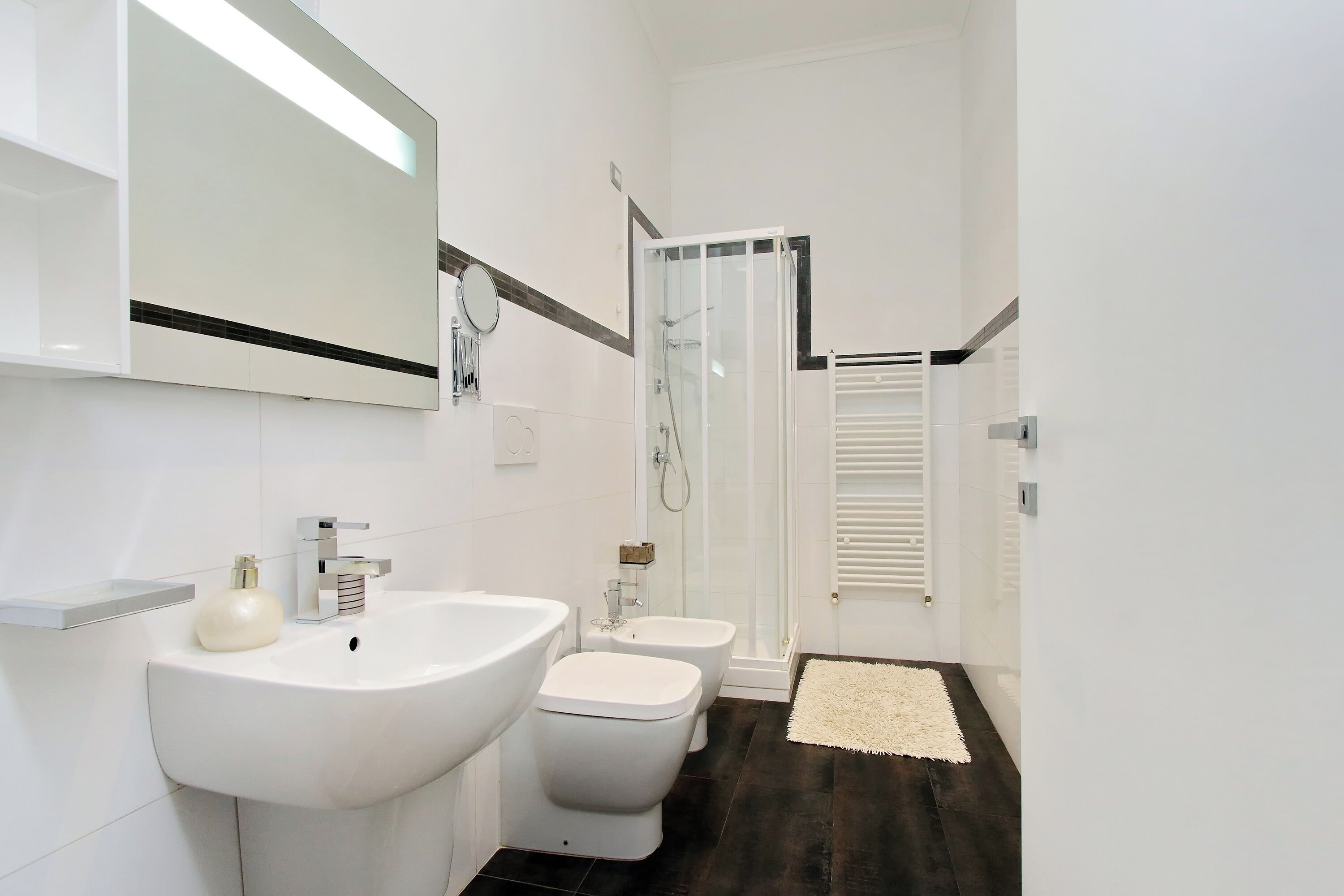 A modern bathroom with a large sink, two toilets, a glass shower enclosure, and a white rug.