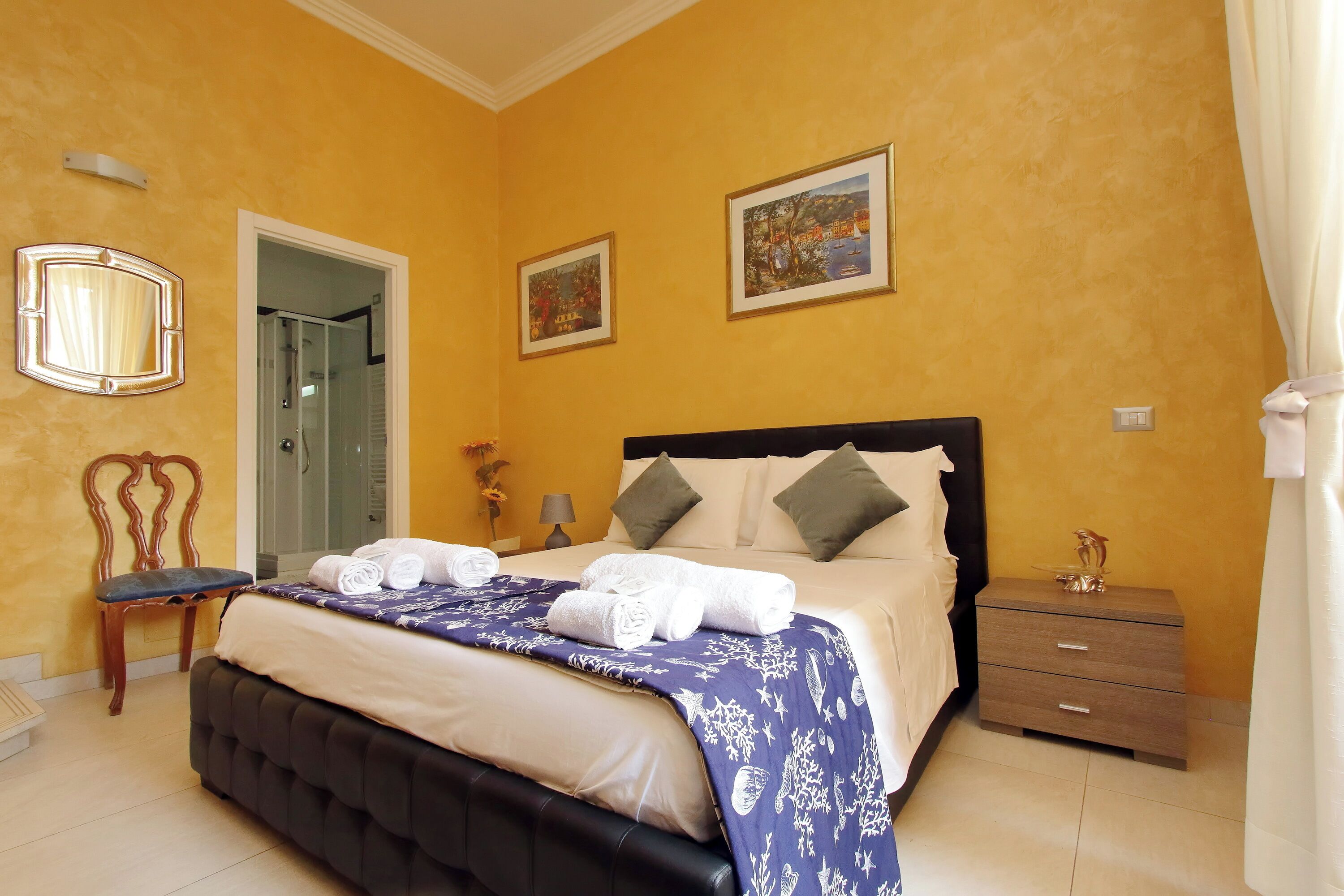 A neatly made bed with a blue and white patterned bedspread, a wooden chair, a bedside table with a lamp, and two framed pictures on the wall.