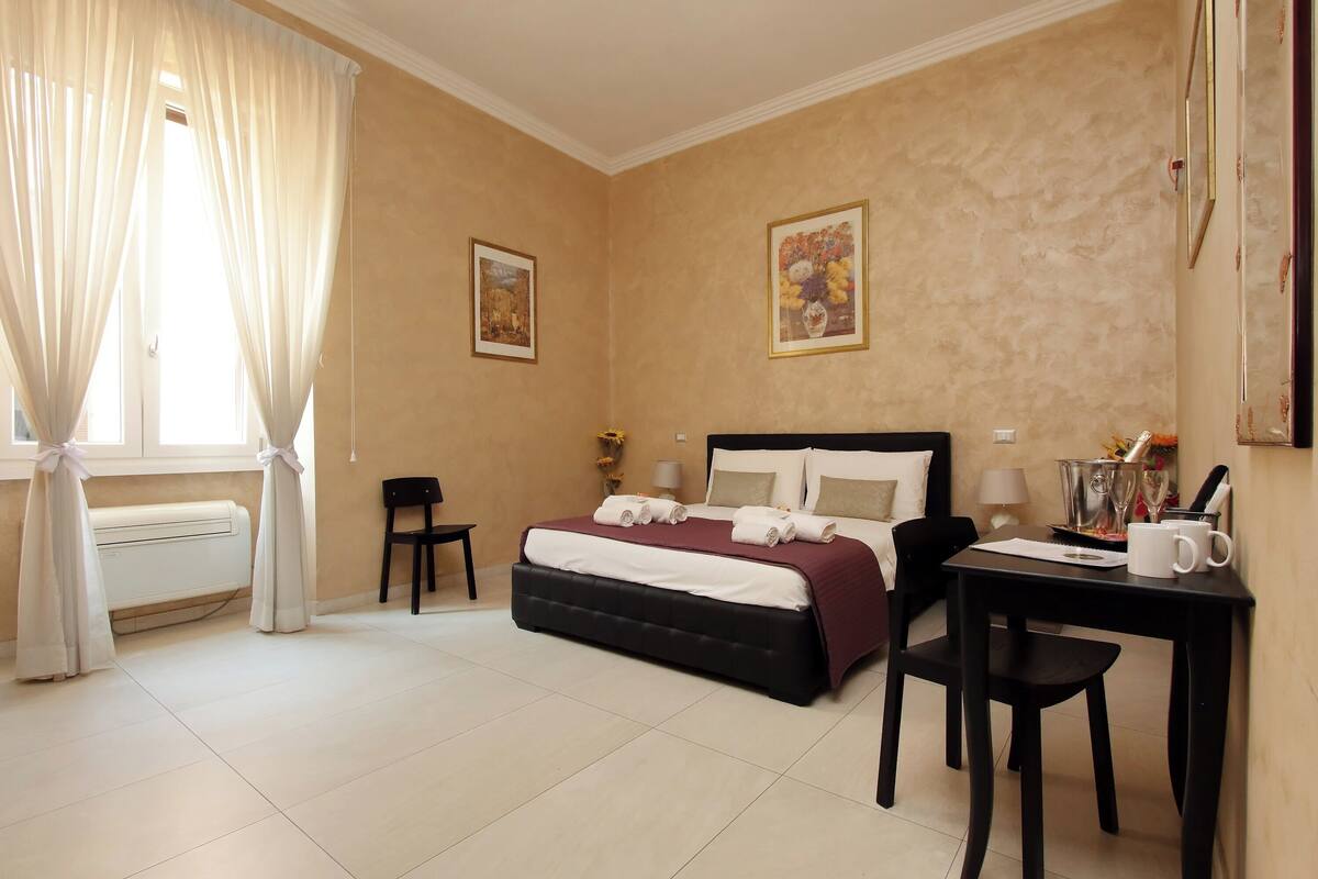 A bedroom with a bed, a desk, a chair, a window with curtains, and two framed pictures on the wall.