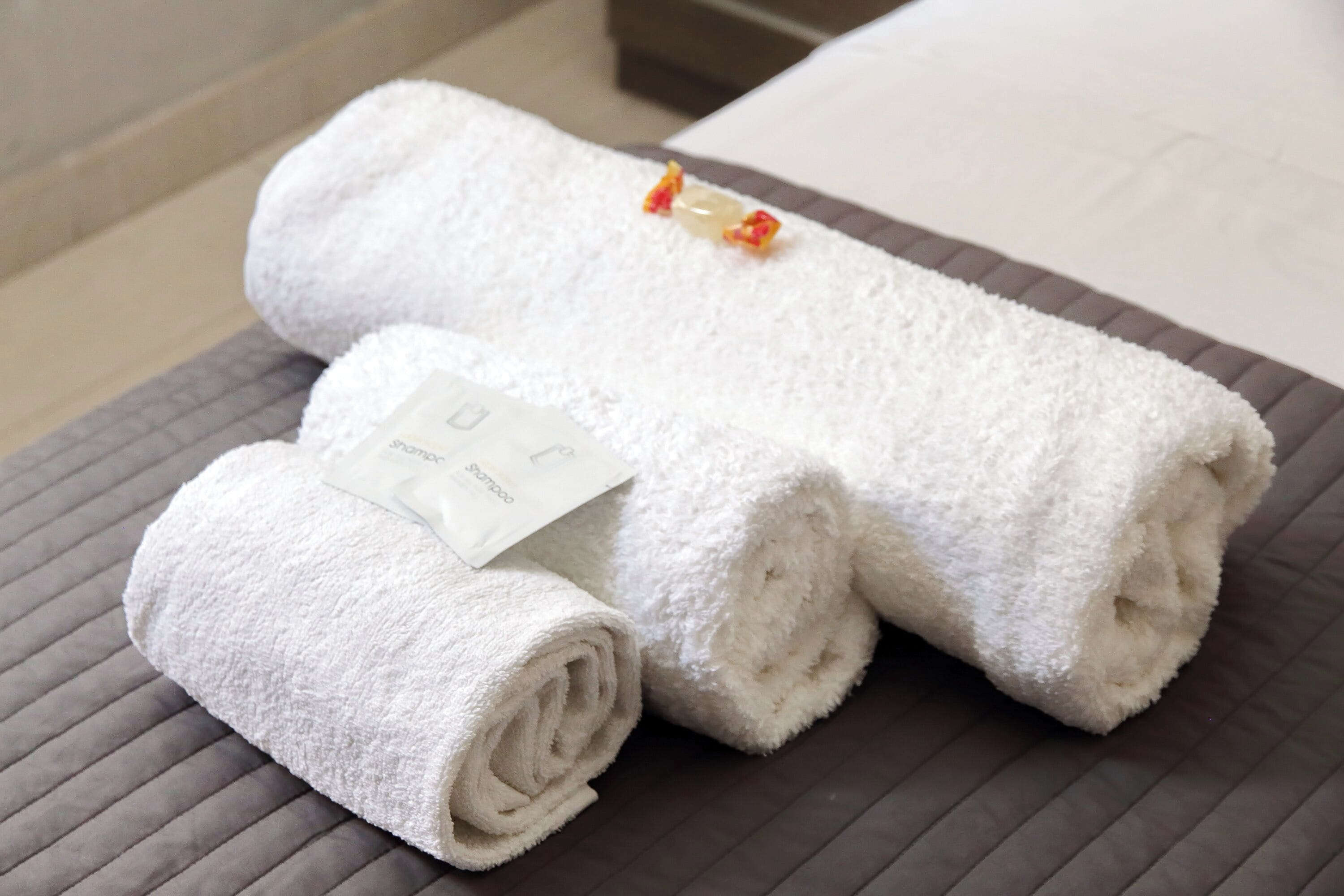 Three rolled white towels on a textured gray surface, likely a bed or seating area.