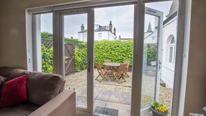 A room with a sliding glass door leading to an outdoor patio area with a table and chairs, a potted plant, and a view of a white building with a church steeple in the background. - Helena Cottage, Elie, Fife (Earlsferry)
