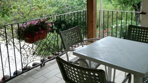 Eira coberta com mesa e cadeiras, rodeada por grades de metal, com vista para um jardim com árvores e flores.