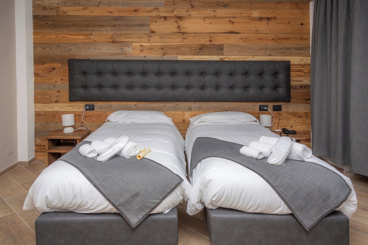 A hotel room with two beds, a wooden wall panel, a grey headboard, and a grey blanket on each bed.