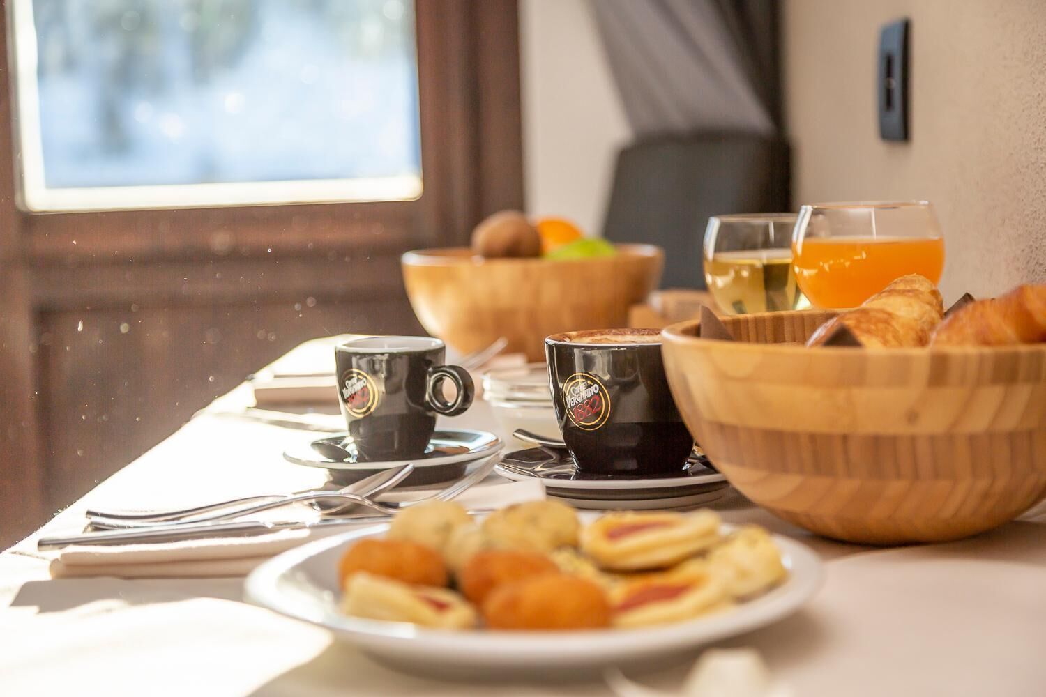 A breakfast setting with coffee cups, a bowl of croissants, and a glass of orange juice on a table.
