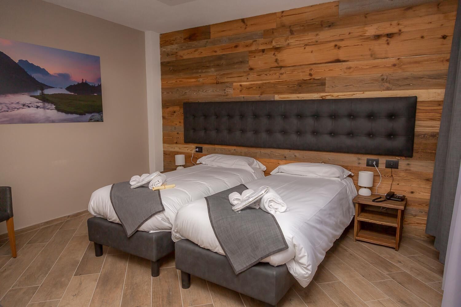 A double room with two beds, a wooden headboard, a painting of a landscape, and a nightstand with a lamp.