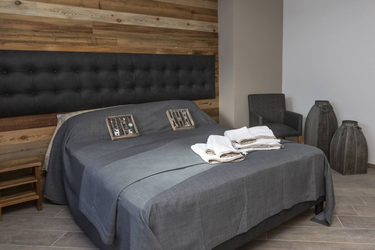 A neatly made bed with a grey bedspread and two pillows, a wooden headboard, a dark armchair, and a tall vase on the floor.