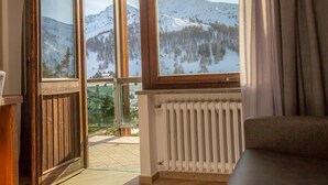 A room with a wooden floor, a sofa, and a view of a snowy mountain landscape through large windows. - Hotel Gran Roc (Sestriere)