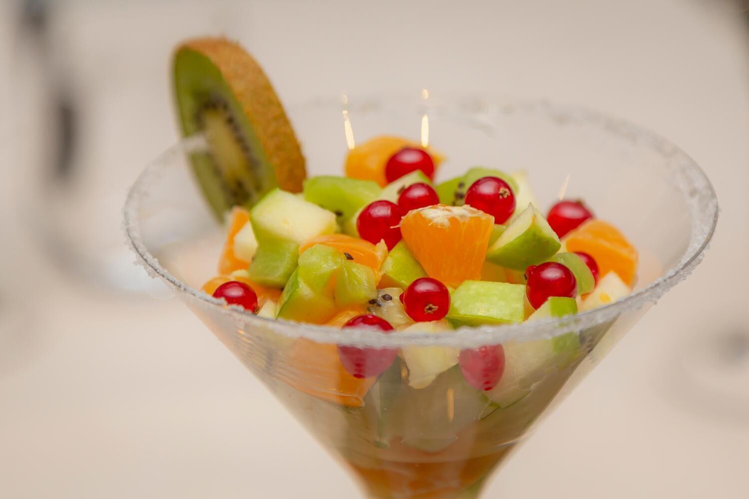 Una ensalada de frutas en un tazón de vidrio transparente, con un trozo de kiwi y una cereza encima.