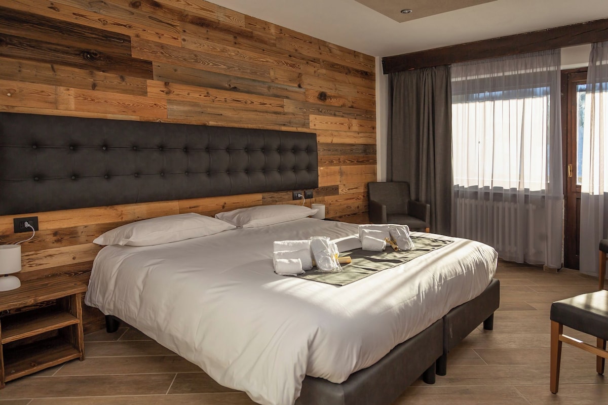 A modern hotel room with a large bed, wooden panel walls, a dark headboard, a chair, and a window with curtains.