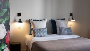 Una cama bien hecha con almohadones estampados, dos lámparas de pared y un cubo de basura pequeño.