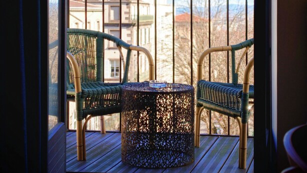 Un balcón con sillones de mimbre y una mesa, con vistas a un paisaje urbano con edificios y cielo despejado.