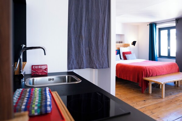 Habitación de hotel compacta con kitchenette, una cama con ropa de cama roja, un banco de madera y una ventana con cortinas azules.