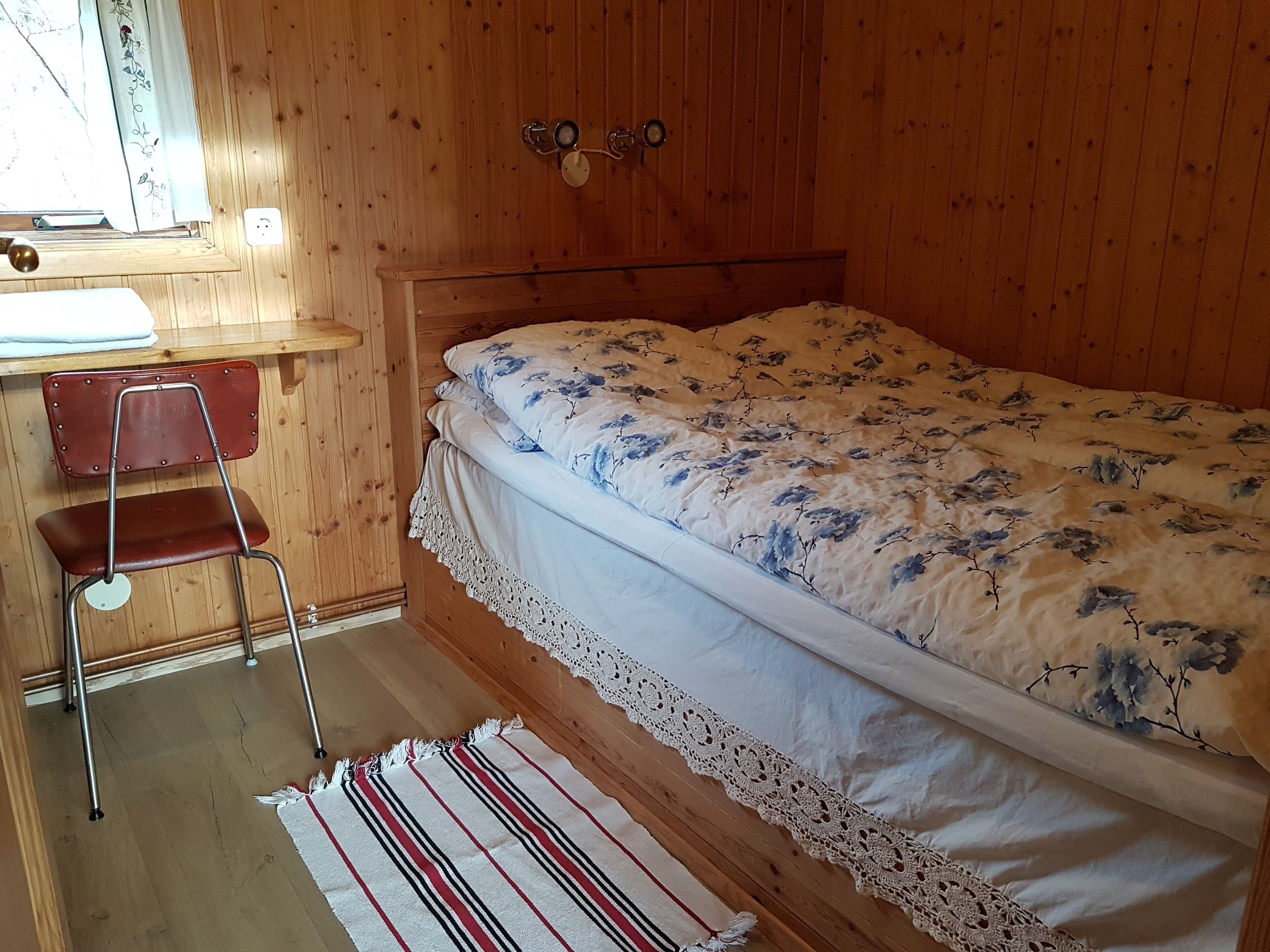 Una habitación de madera con una cama, una silla, una mesita y un ventanal.