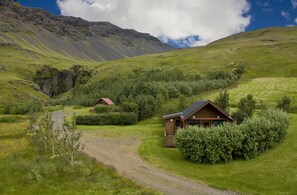 Un chalet rustique, accessible par un sentier de terre, entouré d’une végétation luxuriante et dominé par un paysage montagneux.