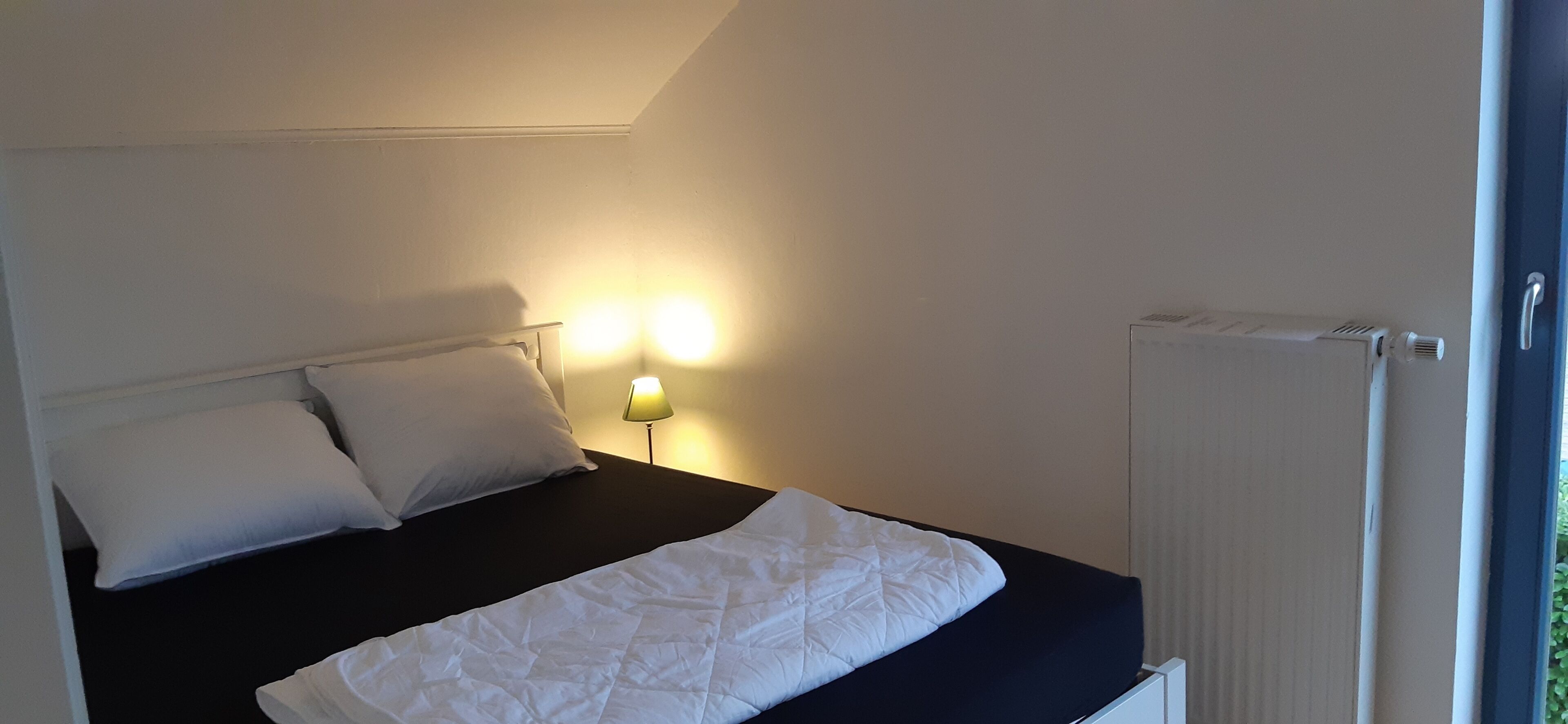 Un letto con biancheria bianca, un comodino con una lampada e un termosifone.
