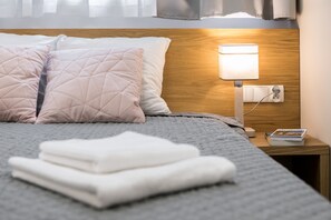 Een netjes opgemaakt bed met een grijze deken, twee roze kussens en een stapel witte handdoeken op een houten nachtkastje.