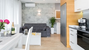 Ein modernes Wohnzimmer mit grauem Sofa, einem Couchtisch und einem Essbereich mit weißem Tisch und Stühlen. Es gibt eine Küchenzeile mit weißen Schränken und einem schwarzen Kühlschrank.