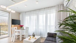 Ein modernes Wohnzimmer mit einer Couch, einem Esstisch mit Stühlen, einem Fernseher und einem großen Fenster mit Vorhängen.