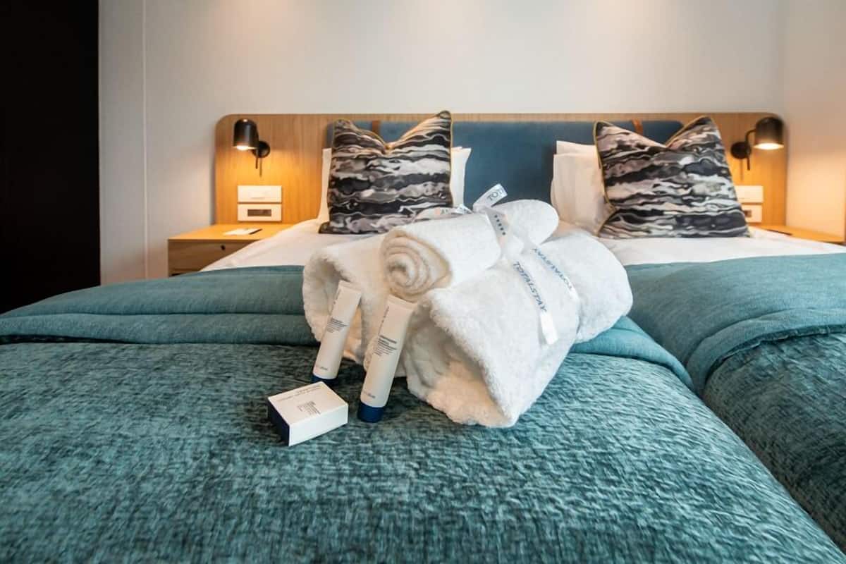 A neatly made bed with a blue bedspread, two pillows with a marbled pattern, and a rolled white towel with a tag.