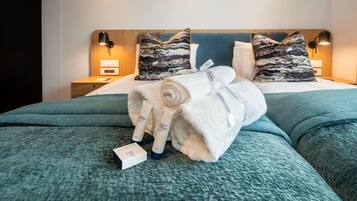 Una cama bien hecha con una colcha azul, dos almohadas con diseño marmolado y una toalla blanca enrollada con etiqueta.