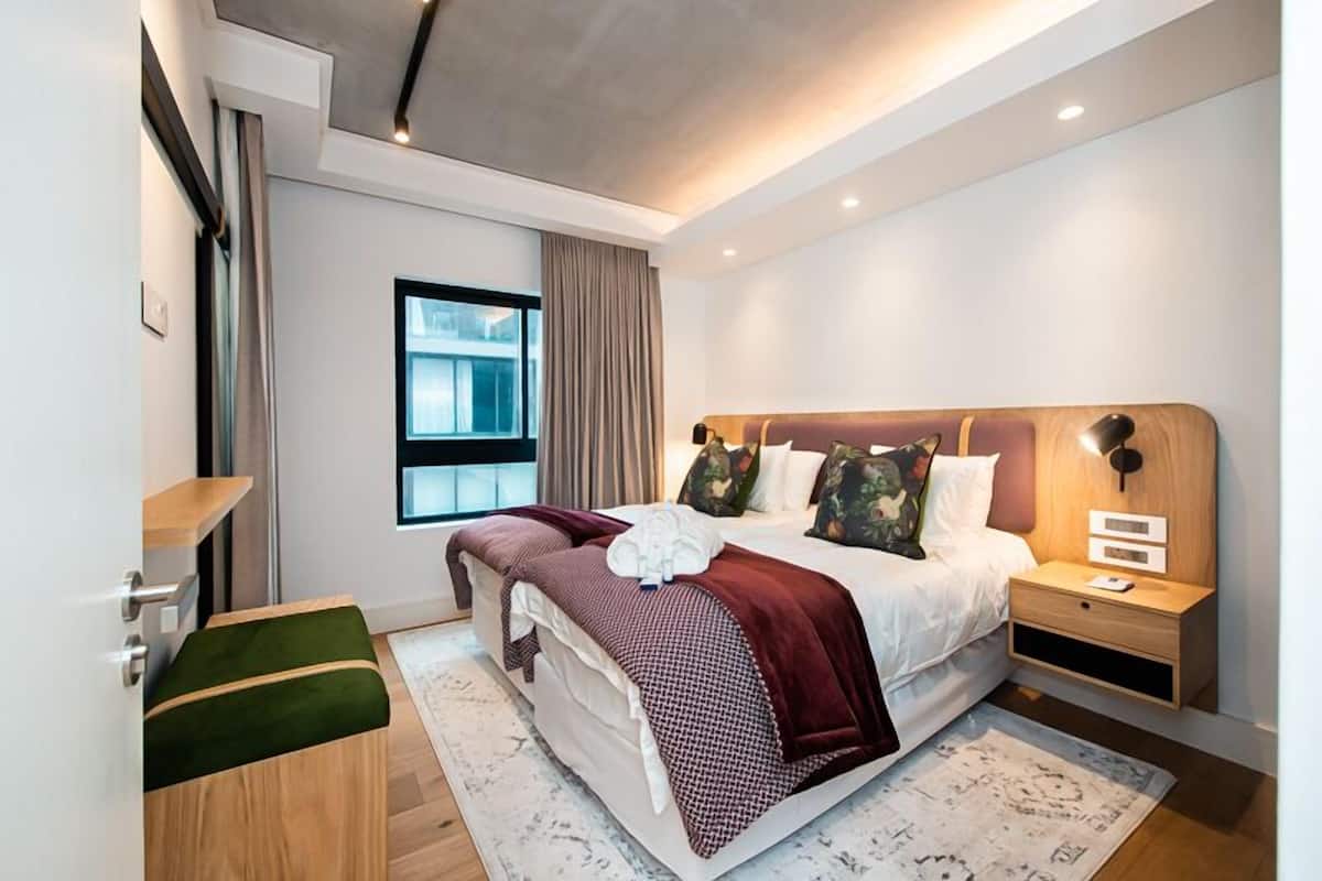A modern bedroom with a large bed, bedside tables, a window, and a view of the city.